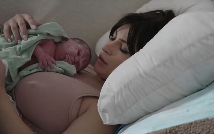 Woman giving birth on Twitch livestream holding newborn baby while resting on a bed with white pillow nearby. Woman giving birth on Twitch livestream holding newborn baby while resting on a bed with white pillow nearby.