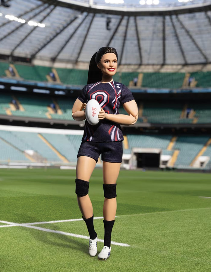 Barbie doll dressed as an Olympic athlete Ilona Maher holding a rugby ball on a green sports field in a large stadium.