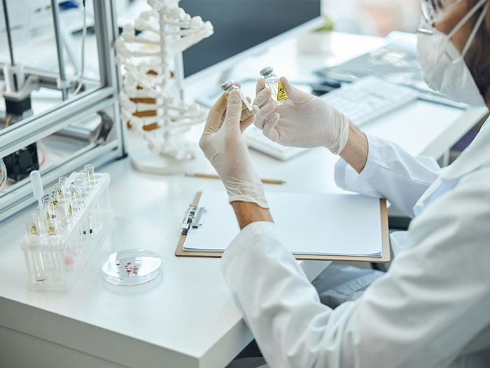 Doctor in a lab wearing gloves and mask, examining vials while reviewing notes on a clipboard next to a skeleton model. Doctor in a lab wearing gloves and mask, examining vials while reviewing notes on a clipboard next to a skeleton model.