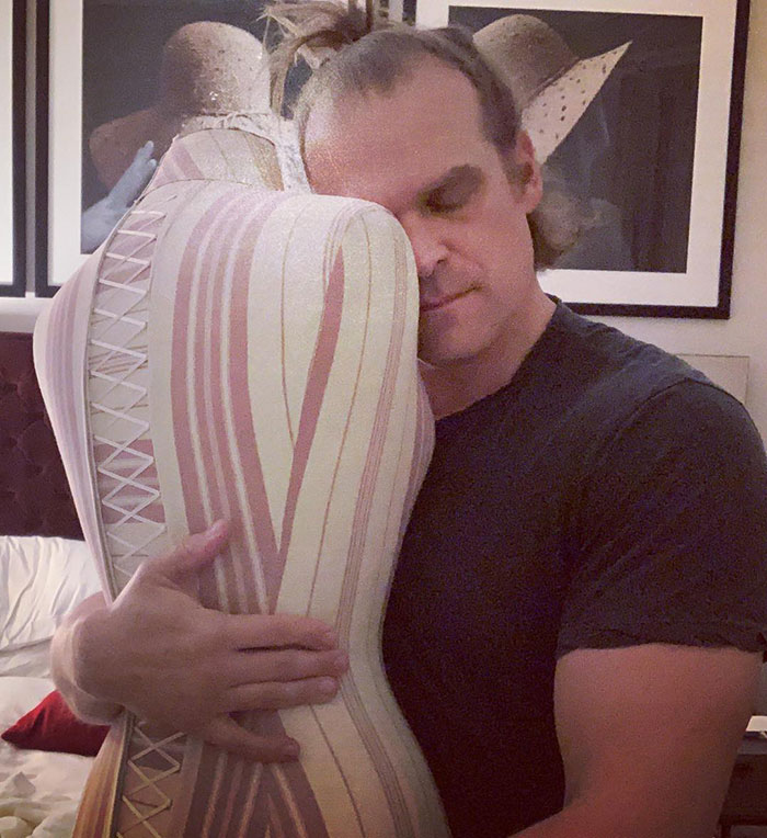 David Harbour embracing a striped mannequin, showing a calm expression in an indoor setting. David Harbour embracing a striped mannequin, showing a calm expression in an indoor setting.