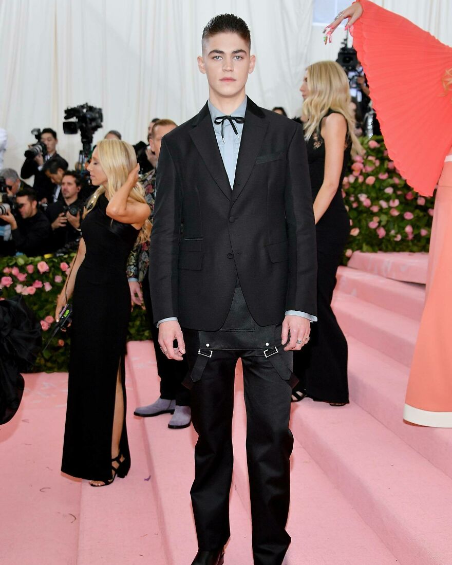 Hero Fiennes celebrity nepo baby wearing a black suit with unique pants posing on a pink carpet at a high-profile event. Hero Fiennes celebrity nepo baby wearing a black suit with unique pants posing on a pink carpet at a high-profile event.