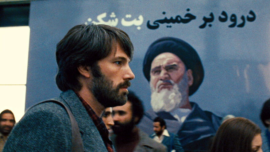 Bearded man walking past a large mural of a religious figure, depicting scenes from movies based on true stories.