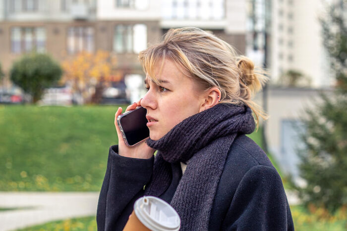 Young woman outdoors using smartphone, holding coffee cup, illustrating genius life hacks that caused extra work realization.