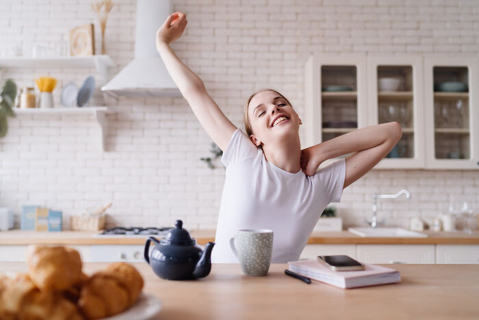 Young woman stretching at kitchen table with tea and pastries, illustrating genius life hacks that reveal extra work tricks.