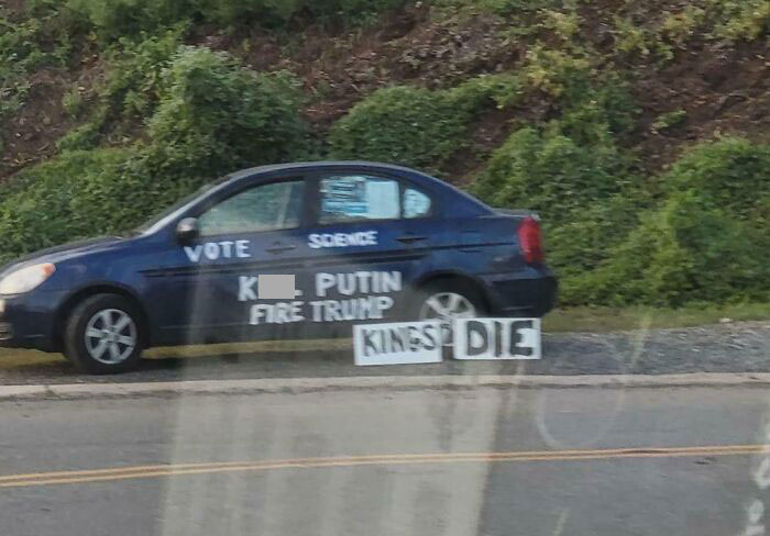 Car with political messages painted on the side and cardboard signs, showing chaotic good expression in public space.