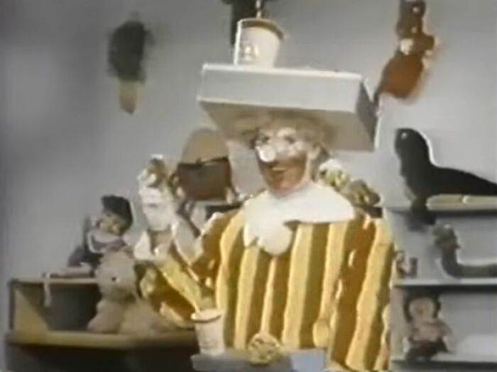 Person in a creepy striped costume holding a cup in a dim room filled with stuffed animals and toys, eerie commercials theme.