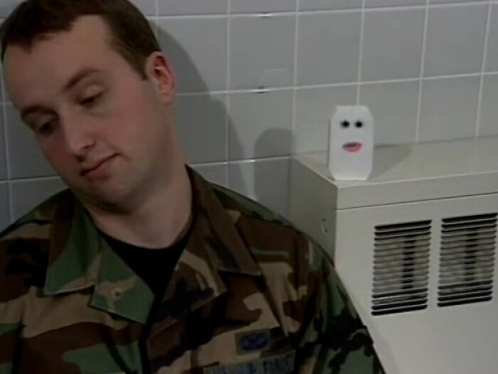 Man in camo uniform leaning with a creepy face on a white object in a tiled room, evoking creepy commercials vibe.