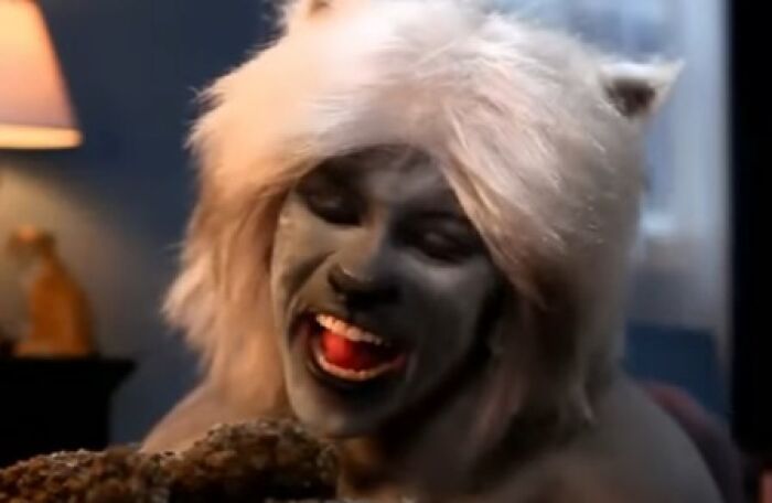 Person in creepy makeup with white hair and dark face paint, smiling eerily in a dimly lit room, from creepy commercials.
