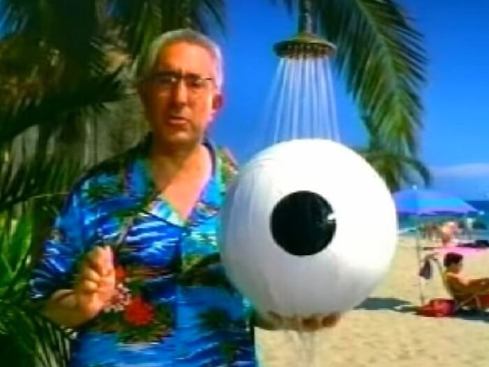 Man in tropical shirt holding a large eyeball prop under a showerhead on a sunny beach, creepy commercials theme.