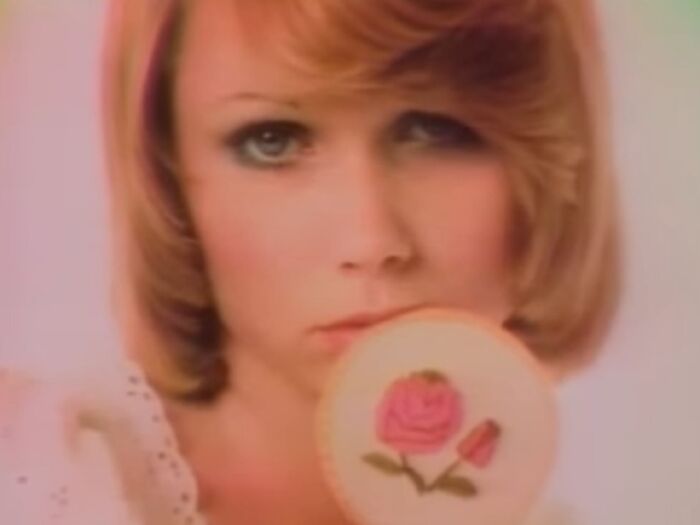 Close-up of a woman with red hair holding a cookie with a pink rose, evoking creepy commercial vibes and unsettling feelings.