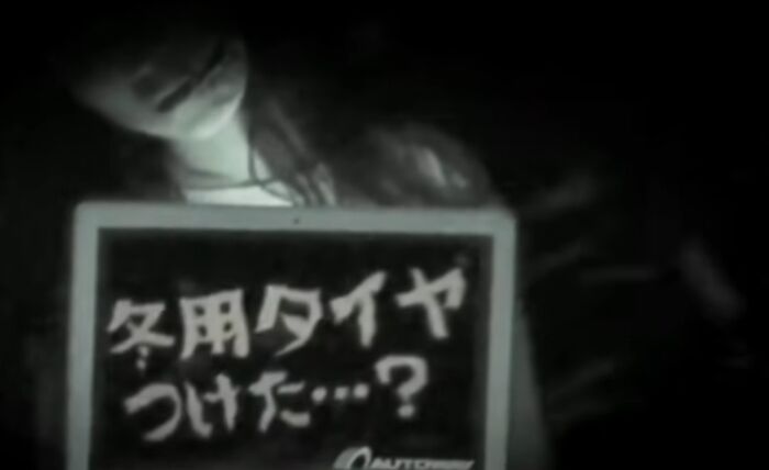 Girl holding a dark sign with Japanese text in a black and white scene from a creepy commercial.