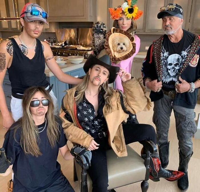 Family dressed in humorous costumes with fake mustaches and a dog in a lion outfit in a kitchen, creating cringed photos.