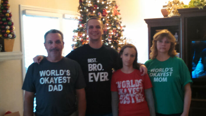 Family photo with four members wearing matching humorous shirts saying world's okayest dad, bro, sister, and mom, funny family photo fail.