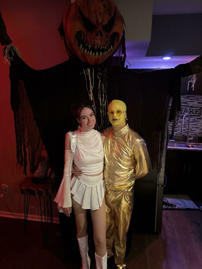 Two people in creative Halloween costumes posing in front of a large spooky pumpkin head decoration at a party.