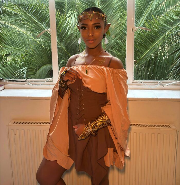 Young woman in a genius Halloween costume wearing a brown corset dress and golden accessories by a window with plants outdoors.