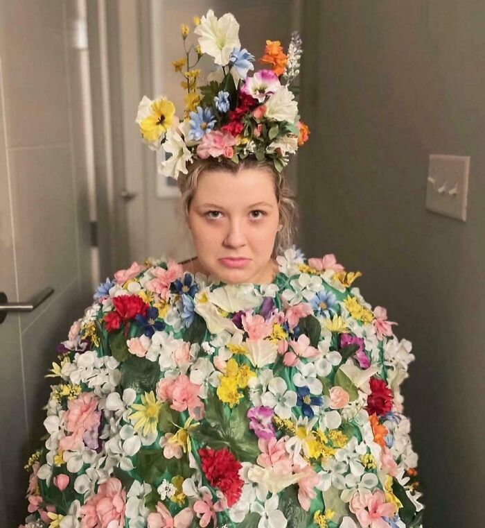 Person wearing a creative Halloween costume covered in colorful artificial flowers and a floral headpiece indoors.
