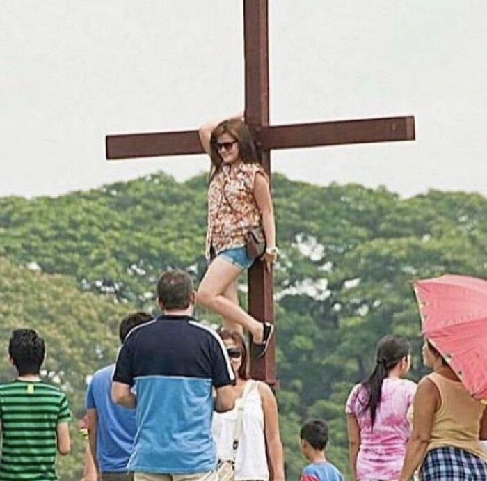 Woman posing humorously on large wooden cross outdoors with a crowd nearby in a funny pleasures of the occult moment.