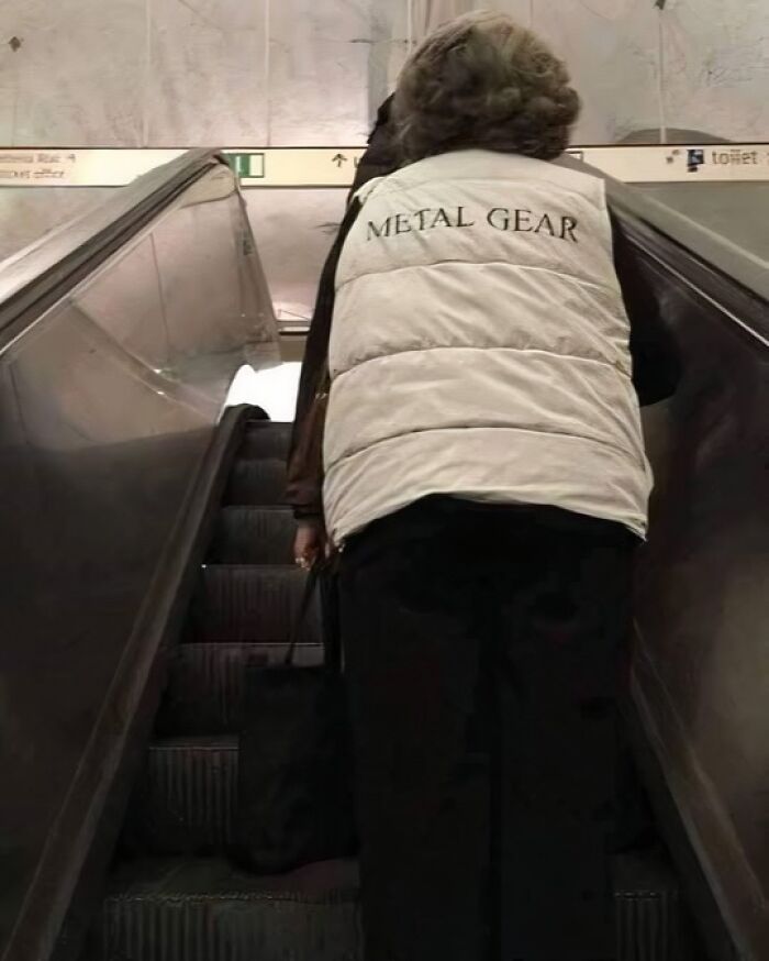 Person wearing a white vest with metal gear text on an escalator, captured in a humorous out of context moment.
