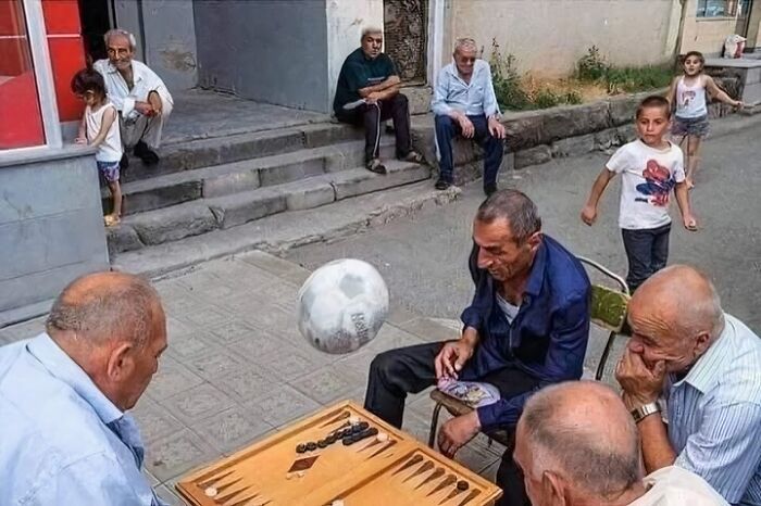 Group of older men seated outdoors playing backgammon while a soccer ball floats nearby in an out of context pleasures of the occult moment