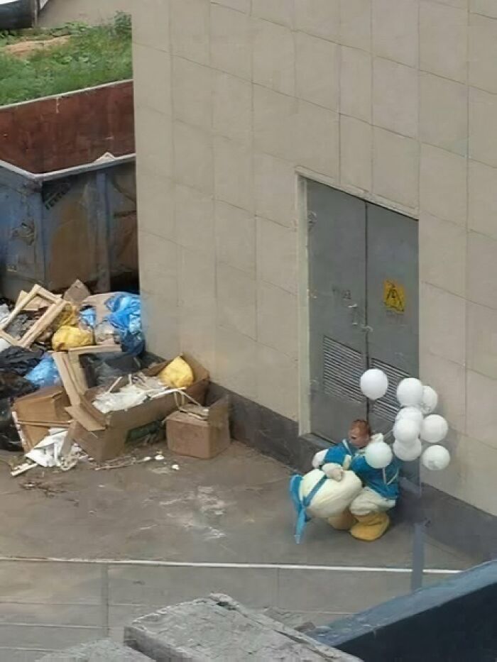 Person in a duck costume crouching near a building door surrounded by trash bags and scattered debris in an outdoor alley.