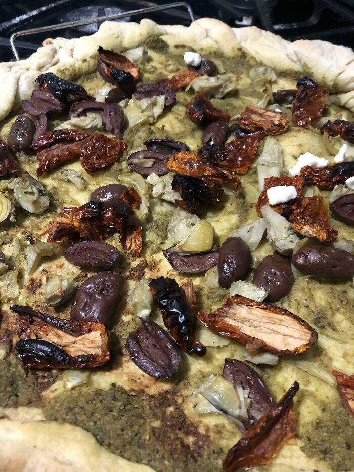 Pie topped with dried olives, charred sun-dried tomatoes, and artichokes on a thick crust, a cursed and disgusting food picture.