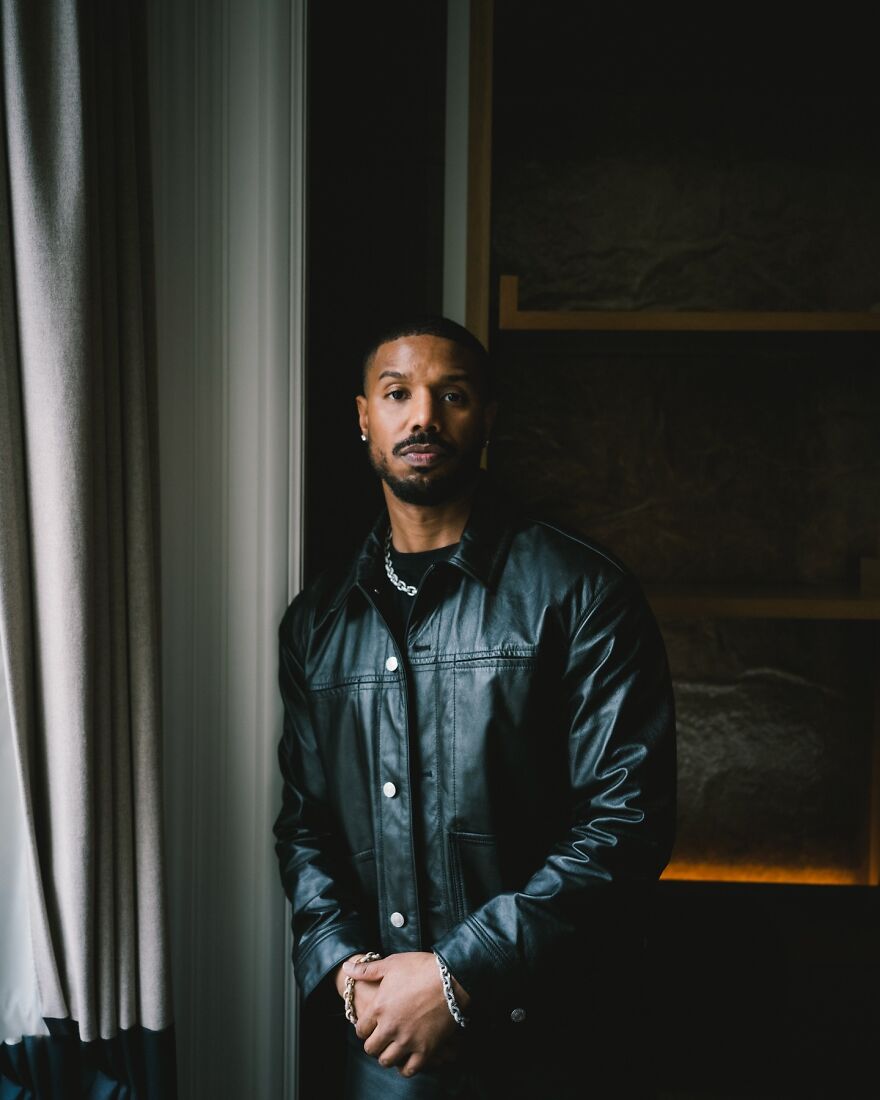 Michael B. Jordan in black leather jacket standing by a window in a dark room, representing most handsome man in the world 2025.