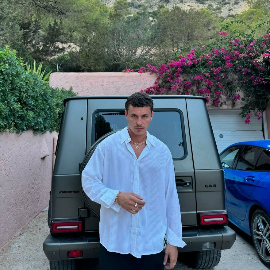 Arón Piper posing in a white shirt with layered gold necklaces near luxury cars, representing most handsome man in the world 2025.