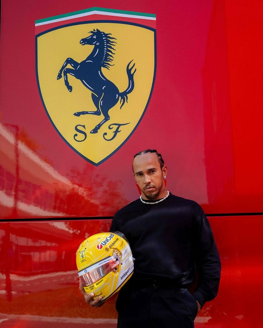 Lewis Hamilton holding a racing helmet standing against a red wall with a large Ferrari logo, representing handsome man 2025.