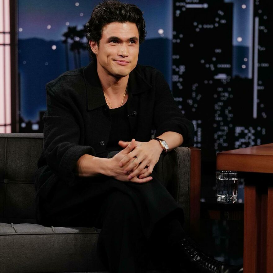 Charles Melton with dark hair and black outfit sitting on a couch during a late-night talk show, representing most handsome man.
