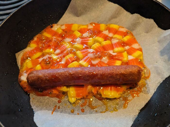 Unappetizing hot dog cooking on melted candy corn in a pan, a cursed and disgusting picture of food.