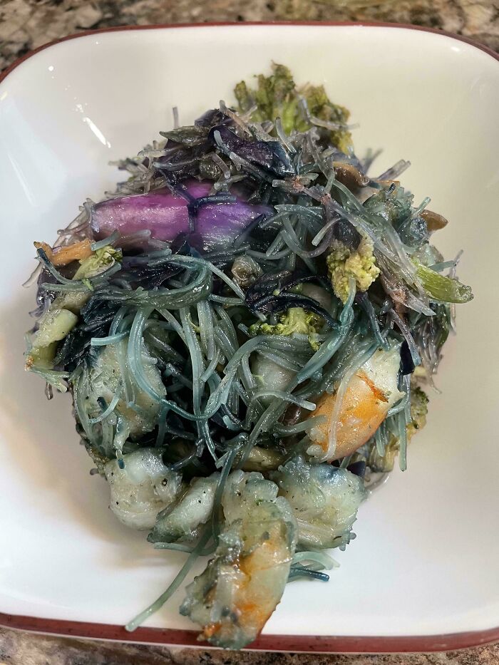 Cursed and disgusting food with slimy noodles, vegetables, and shrimp in a white bowl on a granite countertop.