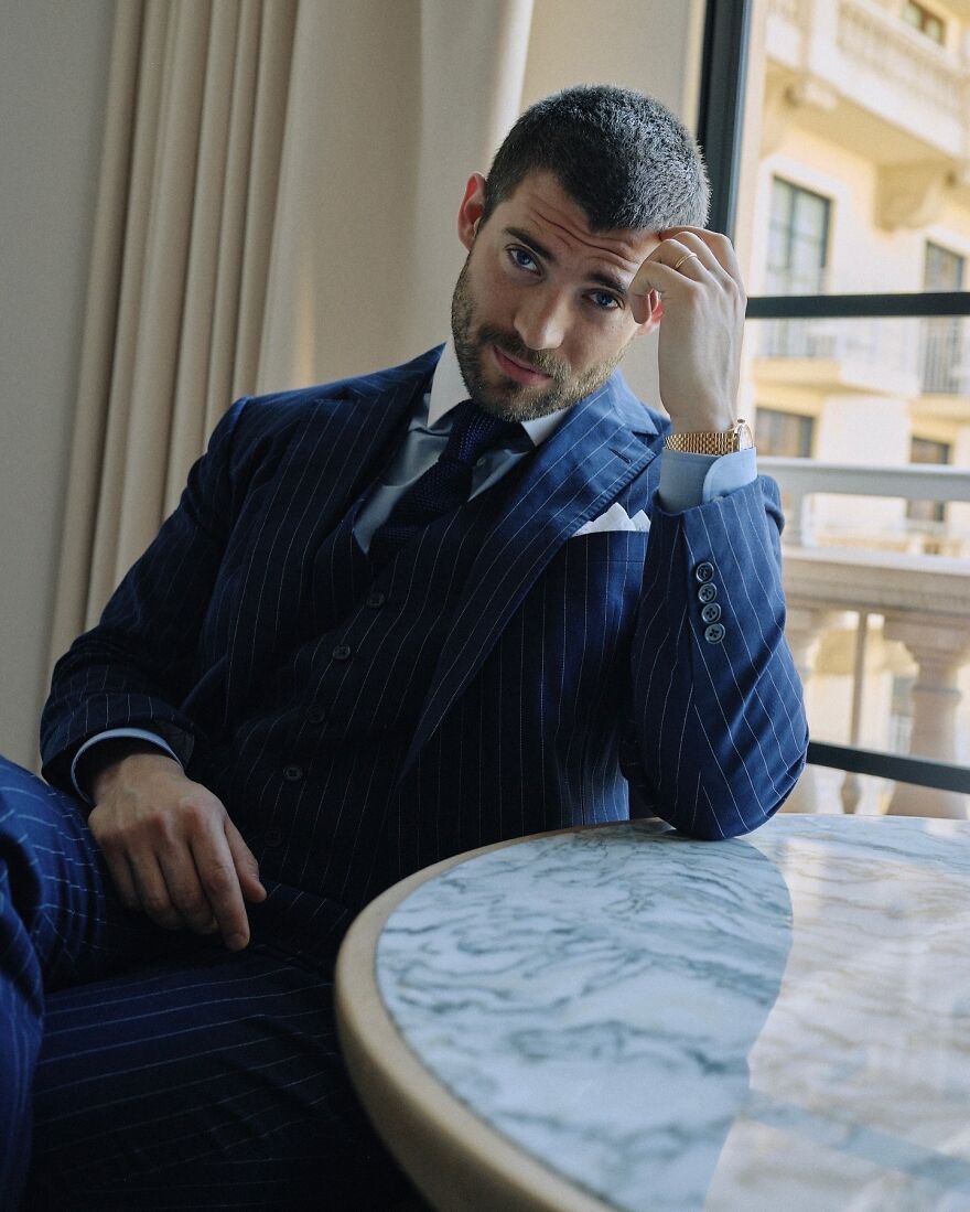 David Corenswet in a navy pinstripe suit sitting by a marble table, posing confidently as a candidate for most handsome man 2025.