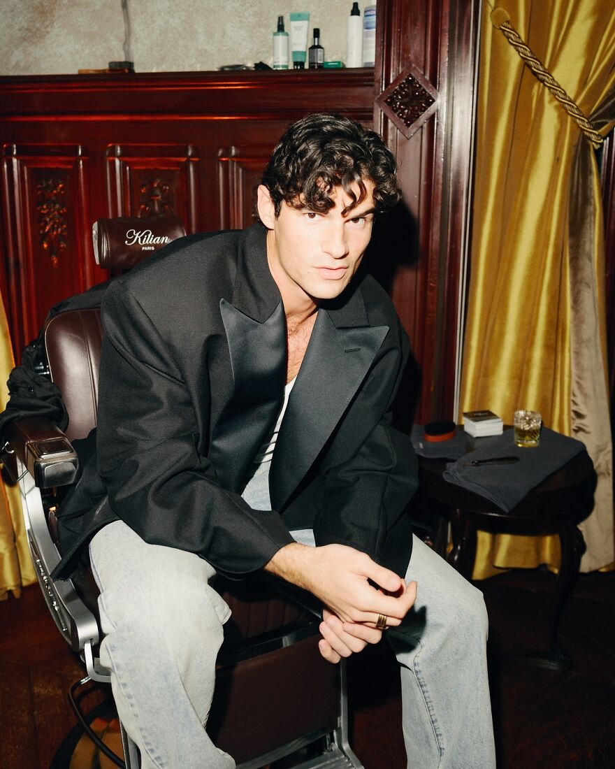 Paul Forman seated indoors wearing a black blazer and jeans, embodying the most handsome man in the world style.