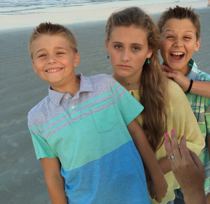 Three kids on the beach with one girl looking unamused and two boys smiling or laughing in a family photo fail.