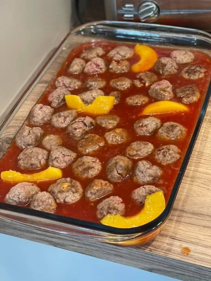 Glass baking dish filled with meatballs and peach slices in red sauce, a cursed and disgusting picture of food.