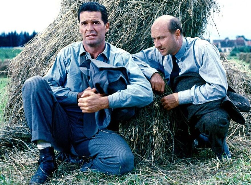 Two men dressed in vintage clothing sitting near a haystack in a scene from movies based on true stories.