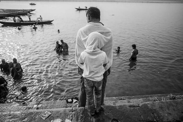 Black-and-white photo of people bathing and boating by the river in the streets of India on stone steps.