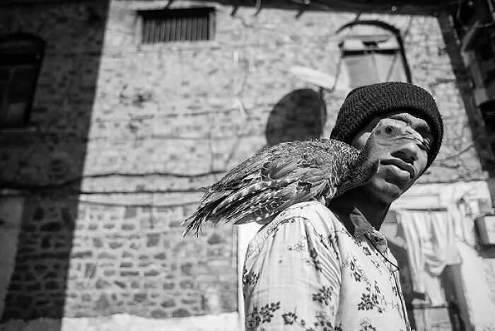 Man wearing a beanie with a bird on his shoulder in a black-and-white photo from the streets of India.