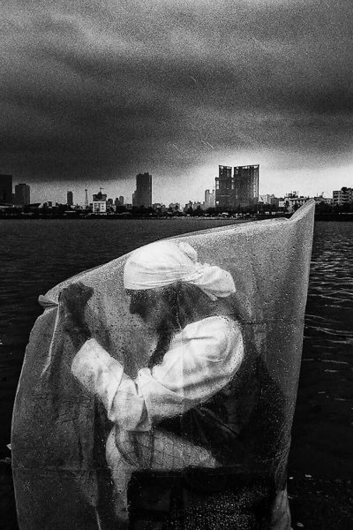 Black-and-white photo of a person in traditional attire covered with plastic near water with a city skyline in the background.