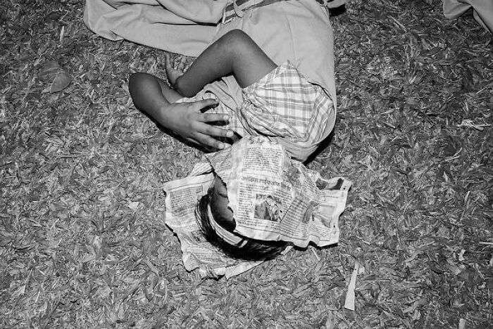 Black-and-white photo of a child lying on grass with a newspaper covering their face on the streets of India.