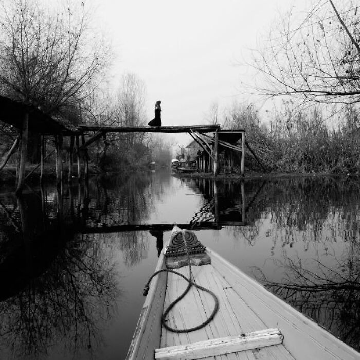 Black-and-white photo of a wooden bridge over a calm river in India with a person walking and a boat in the foreground.