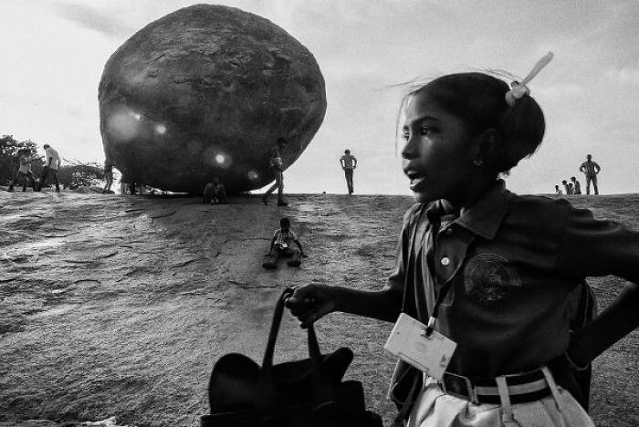 Black-and-white photo showing a girl and people around a large rock on the streets of India at sunset.