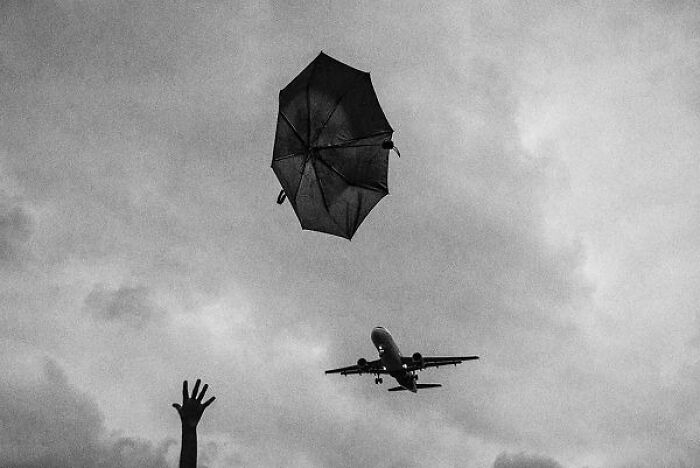 Black-and-white photo from the streets of India showing a hand reaching for a flying umbrella with an airplane overhead.