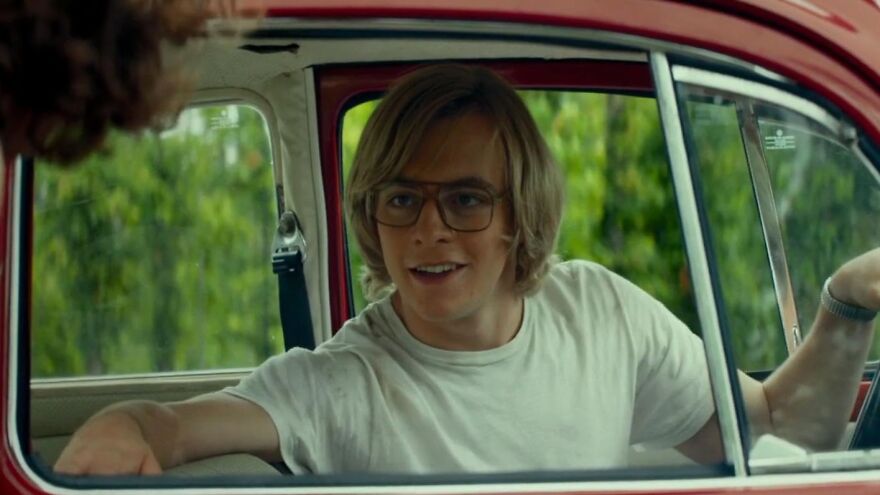 Young man wearing glasses and a white shirt sitting inside a red vintage car in a movie based on true stories.
