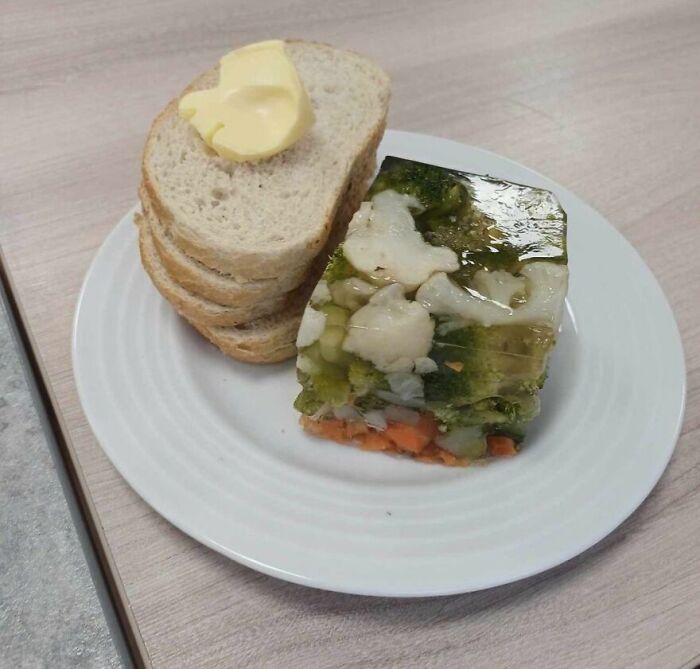 Piece of layered gelatin with vegetables and slices of bread with butter on a white plate, cursed and disgusting food image.