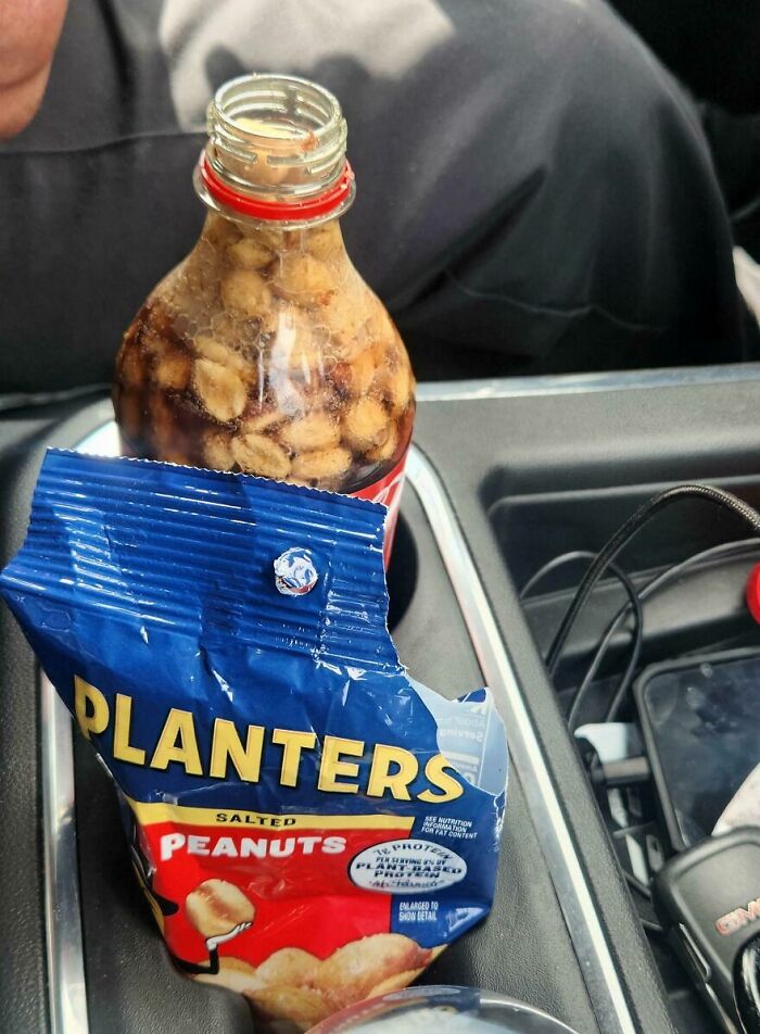 Bottle filled with peanuts soaking inside soda, next to an opened salted Planters peanuts bag in a car cup holder.