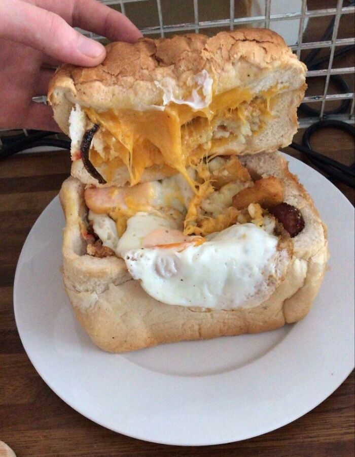 Hand lifting top slice of bread from a cheesy, fried egg and sausage breakfast sandwich on a white plate.