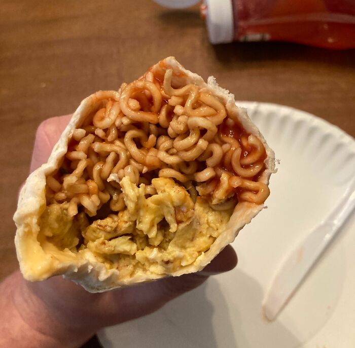 Hand holding a tortilla wrap filled with cooked ramen noodles and scrambled eggs, a cursed and disgusting food combination.
