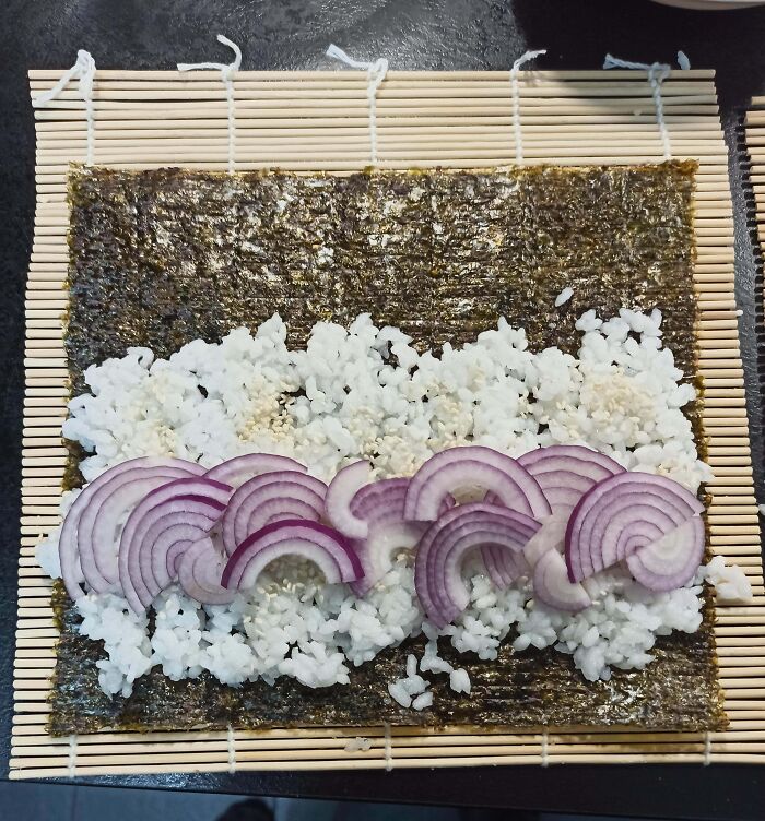 Unappetizing sushi preparation with unevenly spread rice and raw red onion slices on seaweed mat.