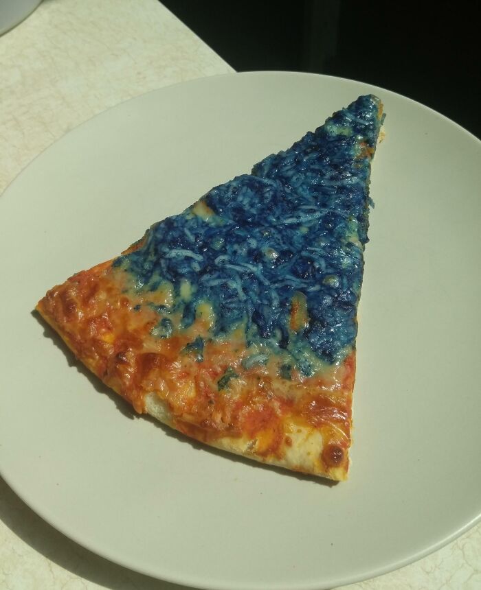 Slice of pizza with blue mold growing on half of the cheese, showing a cursed and disgusting food example on a white plate.
